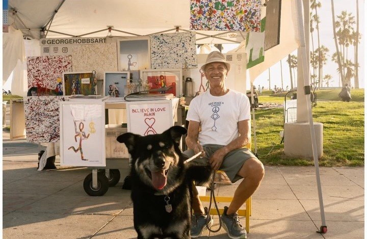 Street artist in a pink hat painting on the Venice boardwalk with his small dog beside him