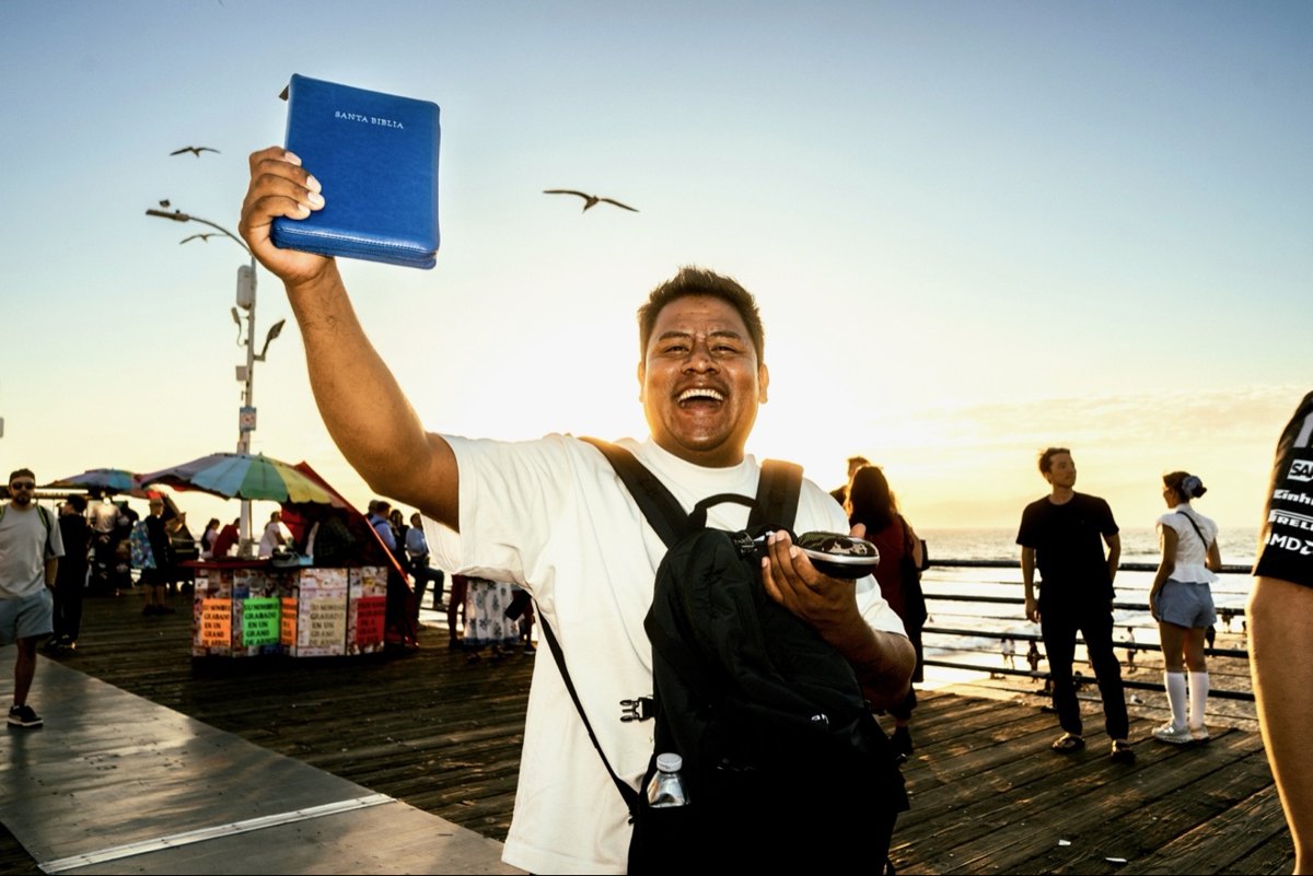 Laughing man on Santa Monica Pier holding up a blue Santa Biblia against the sunset