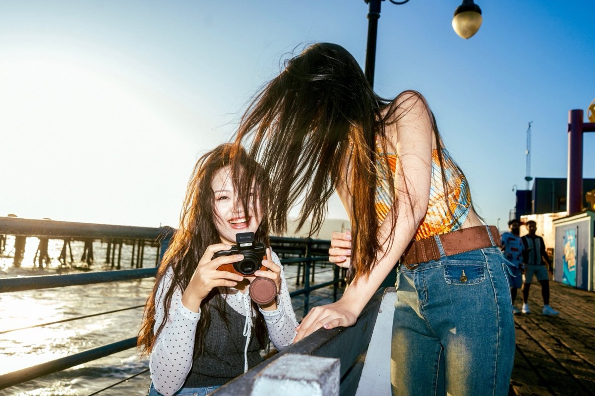 Two friends laughing on Santa Monica Pier, one holding a camera, hair blowing in the wind