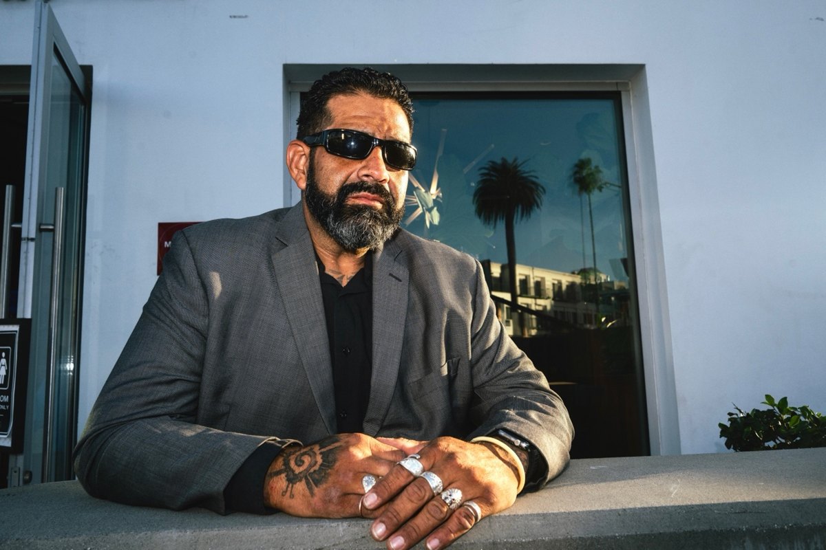 Man in a grey suit and sunglasses with rings and a hand tattoo leaning on a railing
